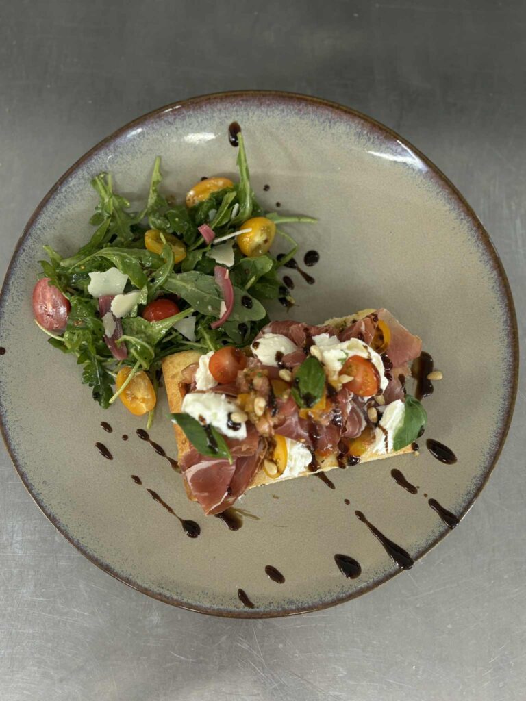 Bruschetta burrata, jambon cru, tomates cerises, basilic et pignons sur toast, garnie de coulis balsamique. Salade roquette, parmesan, oignons.