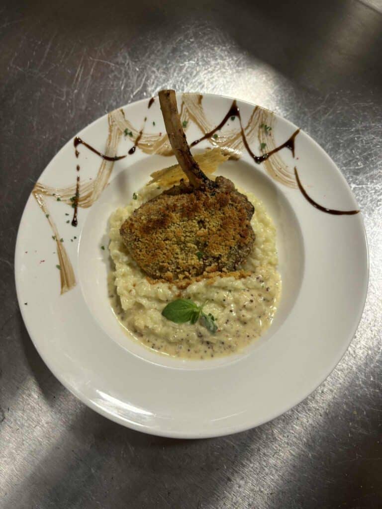 Côtelette panée à l'os sur risotto crémeux, sauce moutarde à l'ancienne et basilic. Dressage élégant sur assiette blanche.
