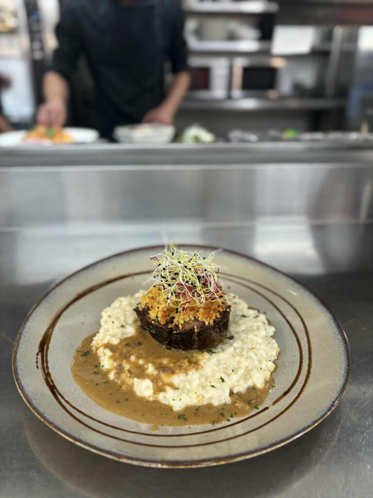 Plat gastronomique : Bœuf en croûte pané sur risotto crémeux, nappé de sauce brune et garni de jeunes pousses. Un chef cuisine en arrière-plan.
