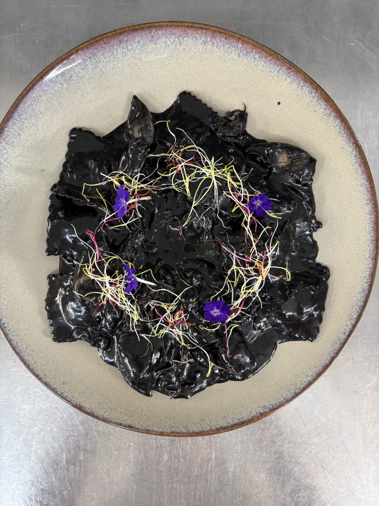 Plat gourmet de raviolis noirs à l'encre de seiche, garnis de pousses vertes et de délicates fleurs violettes sur une assiette ronde.