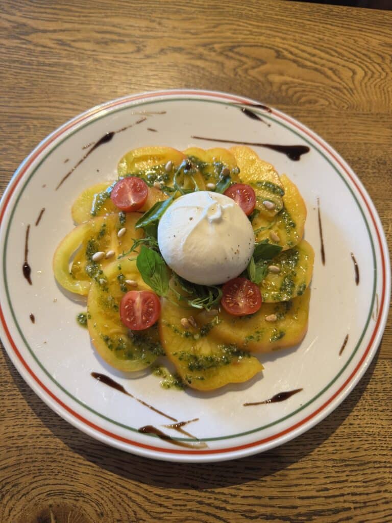 Salade caprese revisitée avec burrata crémeuse, tranches de tomates jaunes, tomates cerises, pesto, basilic et pignons sur une table en bois.