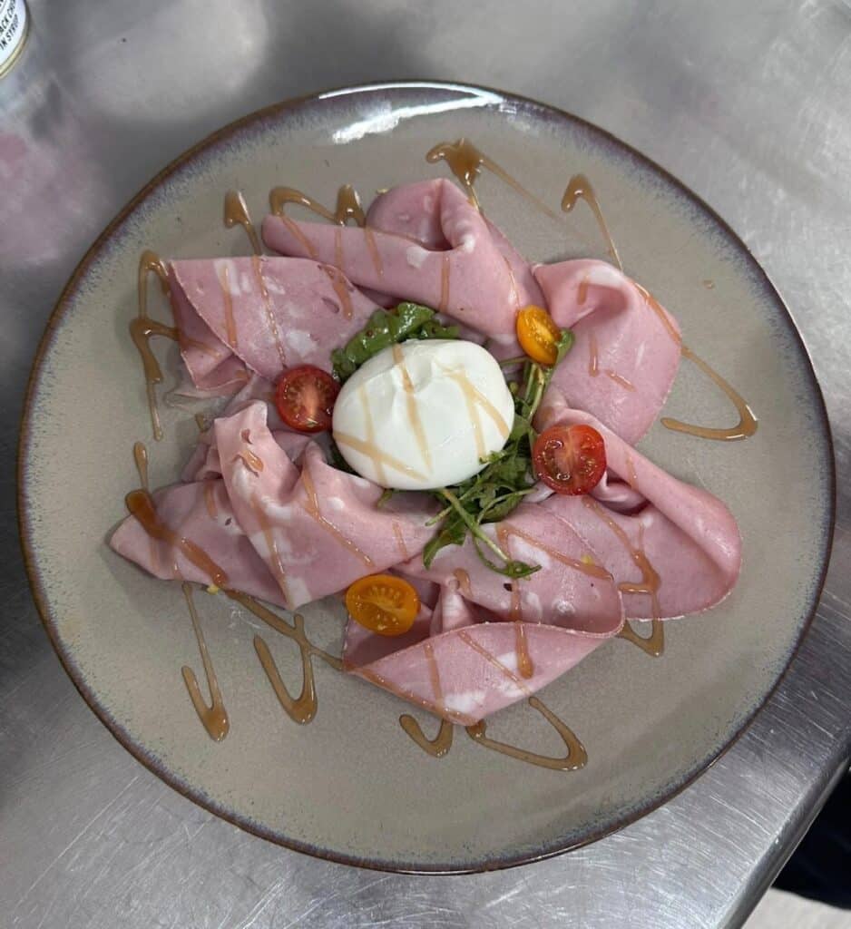 Carpaccio de mortadelle rose, burrata crémeuse, tomates cerises coupées et filet de glaçage sur une assiette grise.