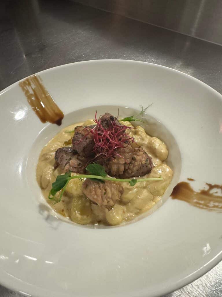 Gnocchis crémeux avec morceaux de viande braisée, garniture de pousses rouges et pois, décorés d'un trait de sauce brune sur assiette blanche.