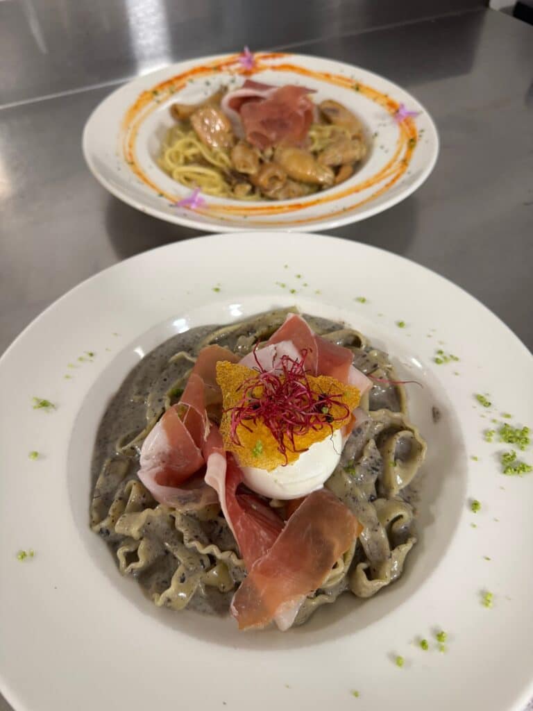 Pâtes tagliatelles foncées aux fruits de mer ou truffe, garnies de tranches de prosciutto, d'un œuf poché et d'une tuile croustillante. Un autre plat de pâtes est flou à l'arrière.