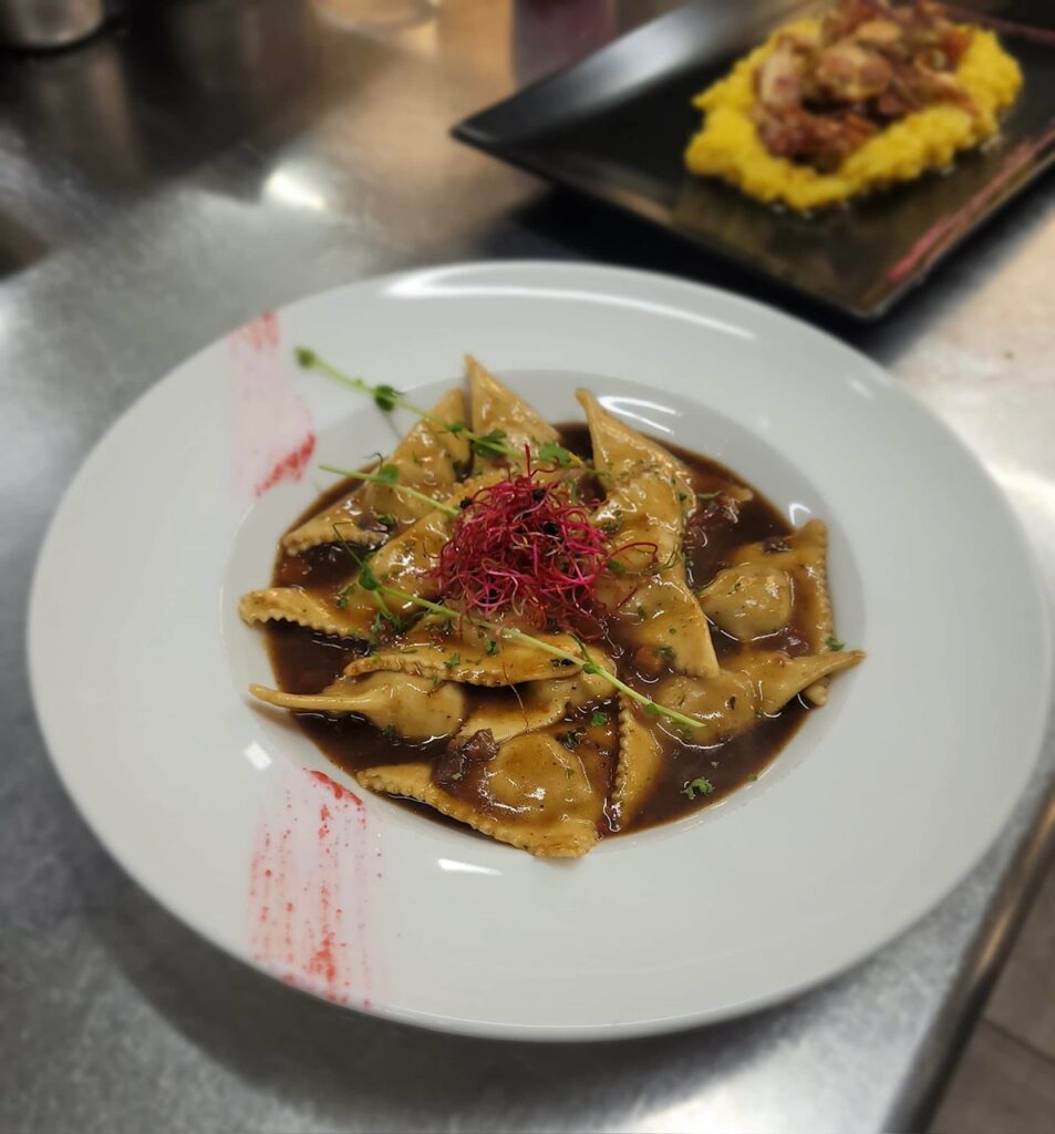 Raviolis triangulaires garnis et nappés d'une sauce brune riche, décorés de micro-pousses rouges et vertes, servis sur une assiette blanche avec une touche artistique rouge.