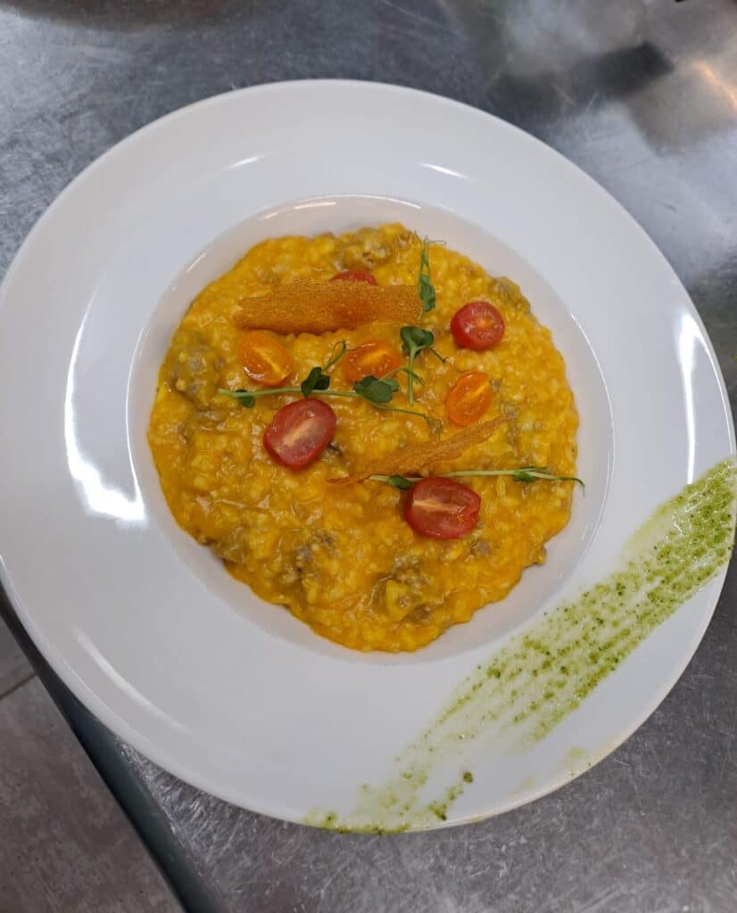 Risotto jaune crémeux avec morceaux de viande, garni de tomates cerises coupées, de pousses et de tuiles croustillantes, sur une assiette blanche décorée de pesto.