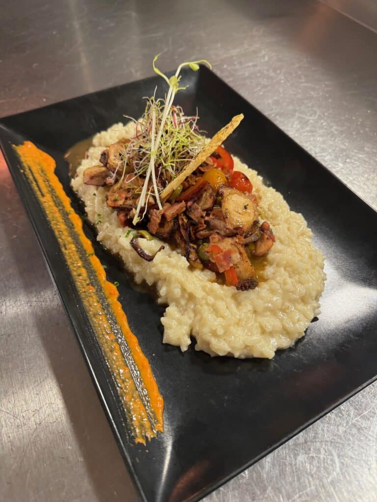 Risotto crémeux sur assiette noire garni de morceaux de poulpe grillé, tomates cerises, micropousses et une rayure de sauce orange vif.
