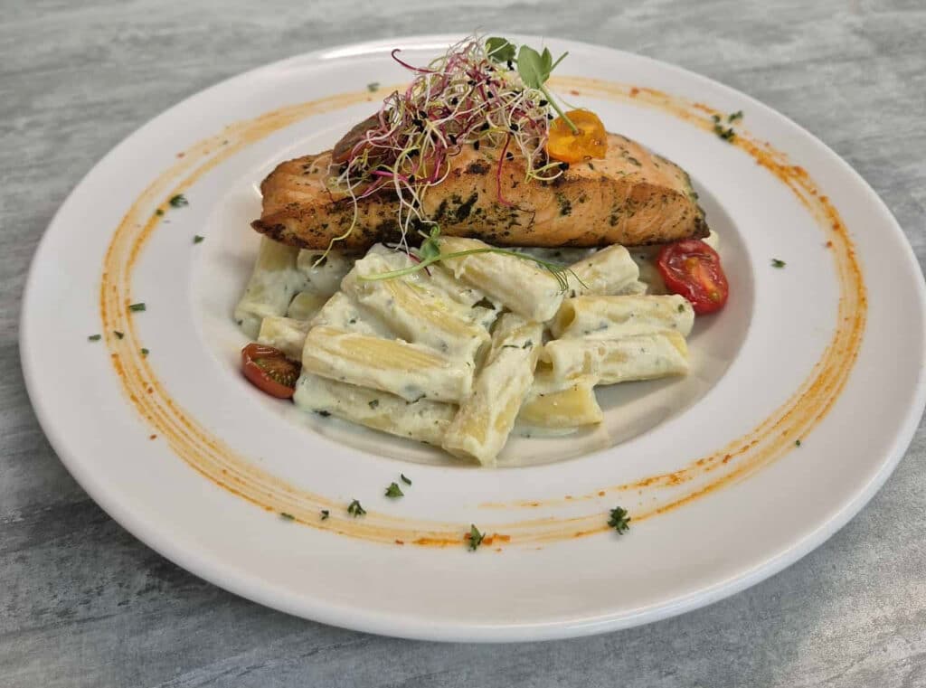 Filet de saumon grillé aux herbes sur des pâtes rigatoni crémeuses, garni de pousses et d'une demi-tomate cerise, présentation soignée.