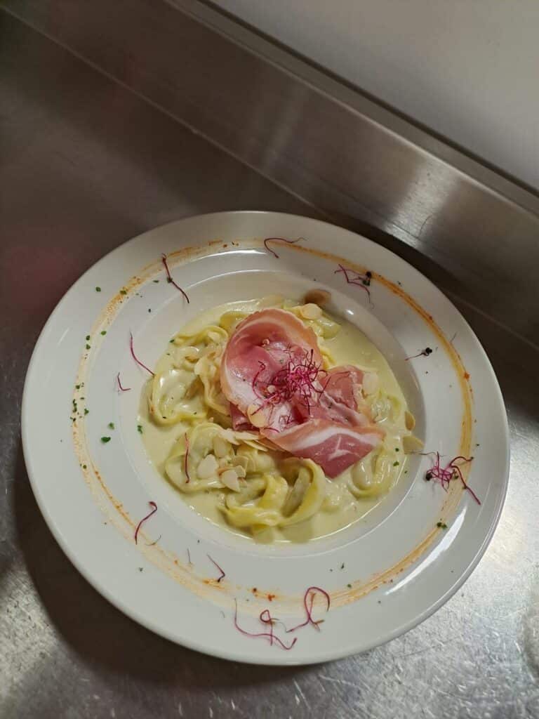 Raviolis tortellinis crémeux, garnis de fines tranches de prosciutto, d'amandes effilées et de jeunes pousses rouges sur une assiette blanche.